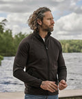 A man with wavy hair and a beard stands outdoors by the water, wearing the Tee Jays Stretch Fleece (9100) zip-up jacket and jeans. Trees and a cloudy sky appear in the background.