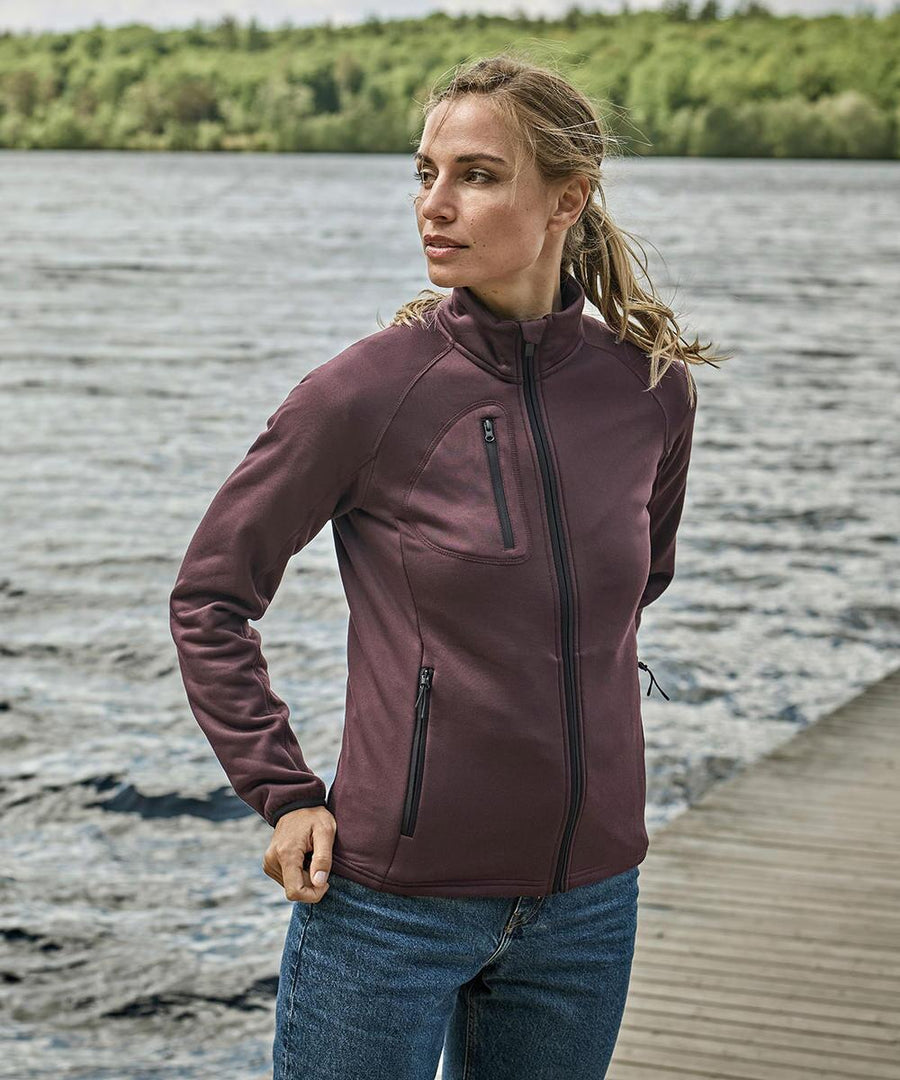 A person wears the Tee Jays Women's stretch fleece (9101) in maroon with blue jeans, standing on a wooden jetty beside water and trees, gazing to the side.