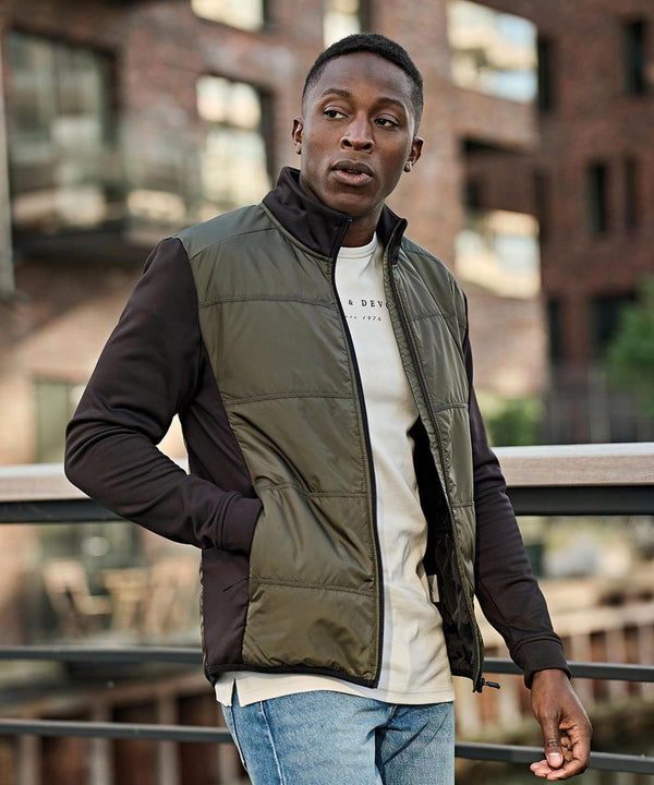 A person stands outdoors in a Tee Jays Hybrid-stretch jacket (9110) over a white shirt and blue jeans, with one hand on a railing and a brick building in the background.