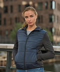 A woman stands outdoors before a blurred brick building, wearing the Tee Jays Women’s hybrid-stretch hooded jacket (9113) in dark blue, hands in her pockets and looking at the camera.