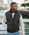 A man in a Tee Jays Hybrid-stretch gilet (9114) over a light grey sweatshirt stands by a riverside railing, gazing left with modern buildings and trees behind him.