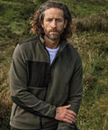 A man with shoulder-length wavy hair and a beard wears the Tee Jays Mountain fleece (9120) in olive green and black over a white shirt, standing outdoors on grassy terrain and looking at the camera with a neutral expression.