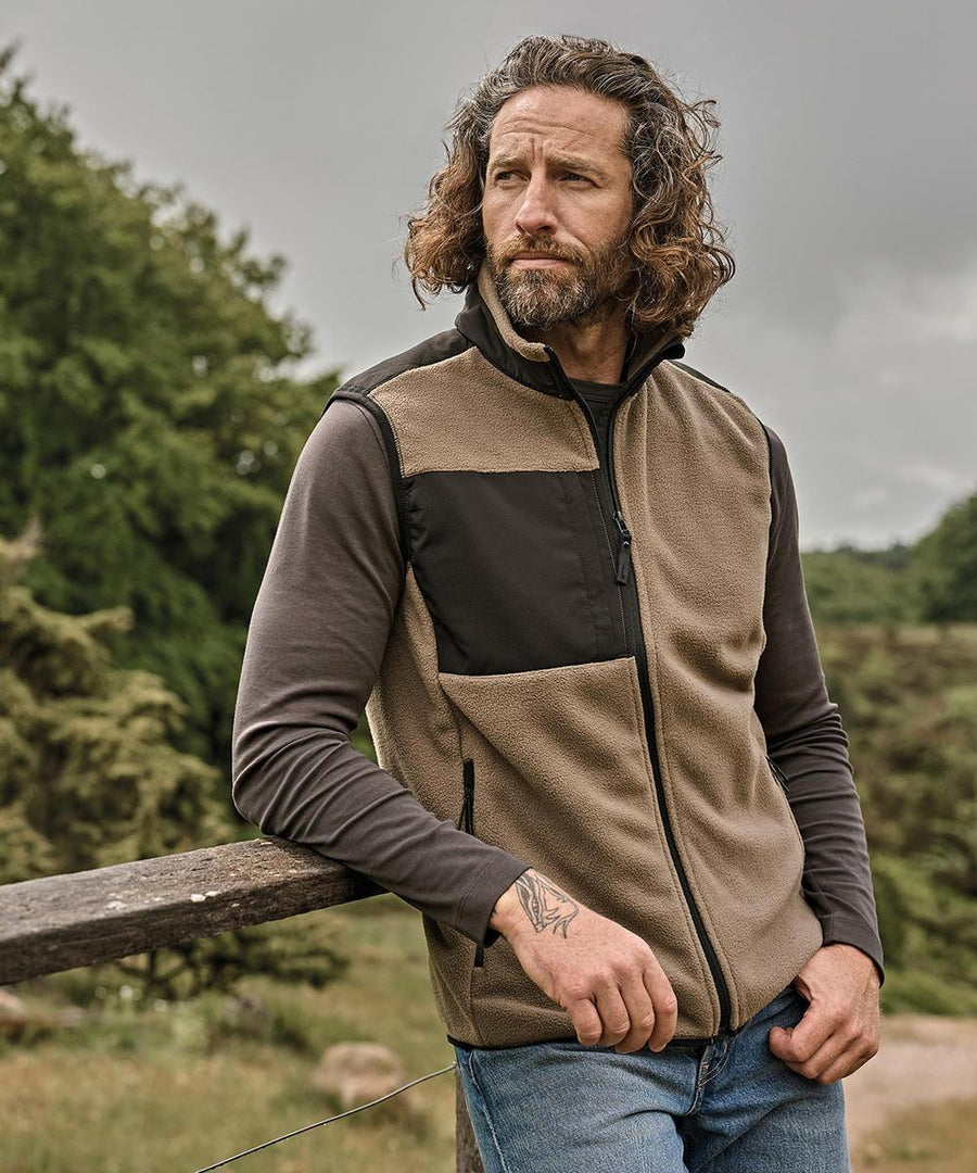 A man with long curly hair and a beard is outdoors, leaning on a wooden fence. He is wearing the Tee Jays Mountain fleece gilet (9122) over a dark long-sleeve shirt and jeans, with trees and greenery in the background.