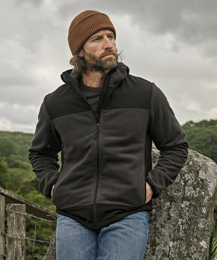 A bearded man wears a brown knitted beanie, blue jeans, and a Tee Jays Mountain hooded fleece (9124) as he stands outdoors by a stone wall and wooden fence with trees and clouds behind him.