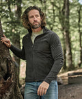 A man with medium-length curly hair and a beard stands outdoors in a wooded area, wearing the Tee Jays Active fleece (9160) over a light shirt and blue jeans, with one hand resting on a tree trunk.