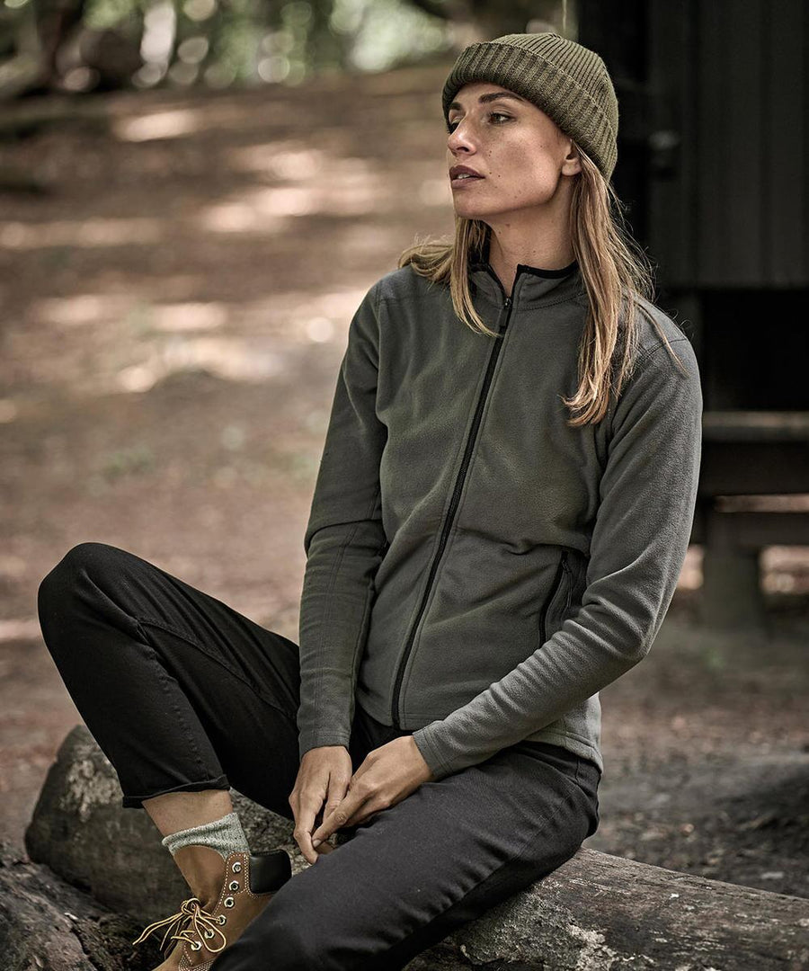 A person with long hair, wearing a green beanie and Tee Jays Women's active fleece (9170), black trousers, and hiking boots, sits on a log in the woods, looking to the side with a neutral expression.
