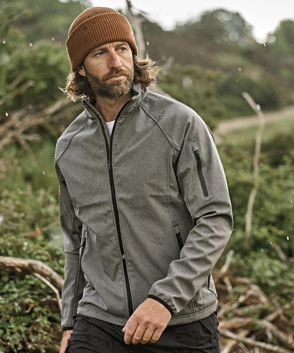 A man with long hair and a beard wears the Tee Jays Lightweight Performance Softshell (9510) in grey and stands outdoors in a natural, green setting, looking off to the side.