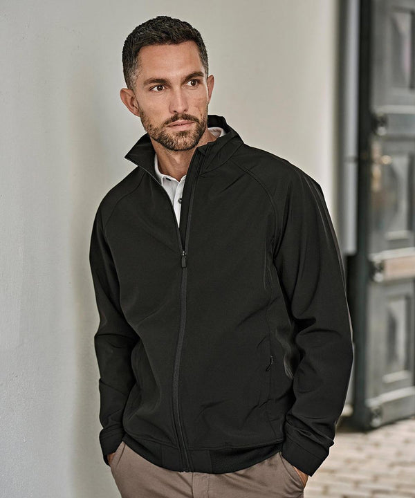 A man with short dark hair and a trimmed beard is wearing the Tee Jays Club jacket (9602) over a light shirt, standing indoors with one hand in his pocket and looking slightly to the side.