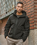 A man with short hair and a trimmed beard wears the Tee Jays Urban Adventure Jacket (9604) in black with beige trousers, standing against a brick wall with one hand in his pocket, looking at the camera.