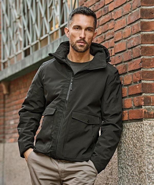 A man with short hair and a trimmed beard wears the Tee Jays Urban Adventure Jacket (9604) in black with beige trousers, standing against a brick wall with one hand in his pocket, looking at the camera.