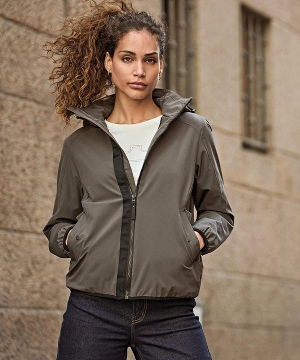 A woman with curly hair stands against a stone wall, wearing the Tee Jays Women’s Urban Adventure Jacket (9605) over a white shirt and dark jeans. She has her hands in the jacket pockets and looks directly at the camera.