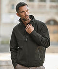 A man with short hair and a beard stands outdoors in a blurred background, wearing the Tee Jays All weather jacket (9606) zipped up and beige trousers, with one hand touching the jacket’s collar.