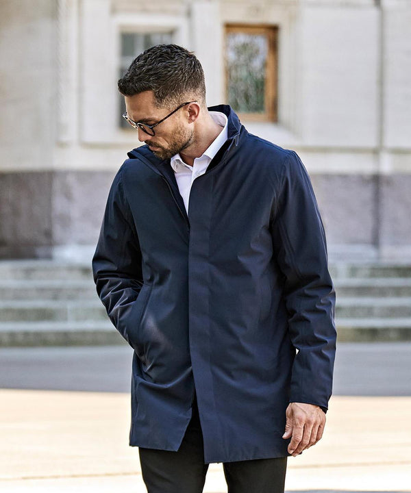 A man wearing glasses, a white shirt, and a navy blue Tee Jays All Weather Parka (9608) stands outdoors with one hand in his pocket, looking down, with stone steps and a building in the background.