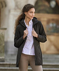 A woman stands outdoors wearing the Tee Jays Women's All Weather Parka (9609) over a white shirt and beige trousers. She holds the parka with both hands, looking left with stone steps and a blurred background behind her.