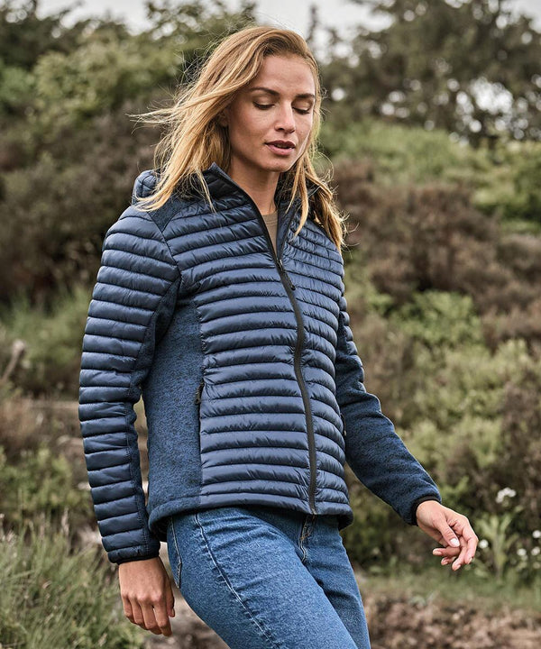 A woman with light brown hair stands outdoors in greenery under an overcast sky, wearing the Tee Jays Women's hooded outdoor crossover (9611) jacket and blue jeans.
