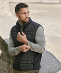 A man with short dark hair and a beard wears a grey polo neck and the Tee Jays Crossover bodywarmer (9624), standing outside by a stone structure and looking to the side.