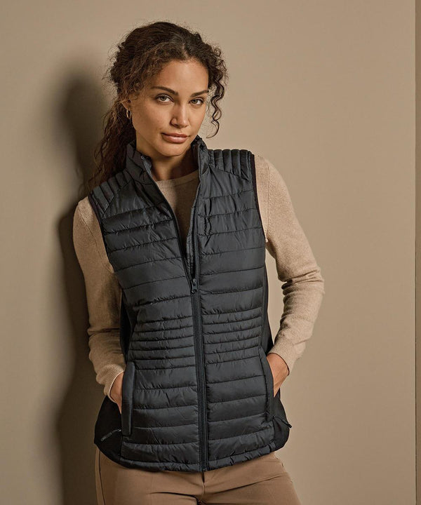 A woman with wavy brown hair stands against a beige wall, wearing tan long-sleeve shirt and trousers, layered with the Tee Jays Women's crossover bodywarmer (9625), her hands in its pockets as she looks at the camera.