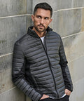 A man with short hair and a trimmed beard wears the grey Tee Jays Crossover jacket (9626) with black sleeves, leans against a stone wall, looking to his left with his hands in his pockets.