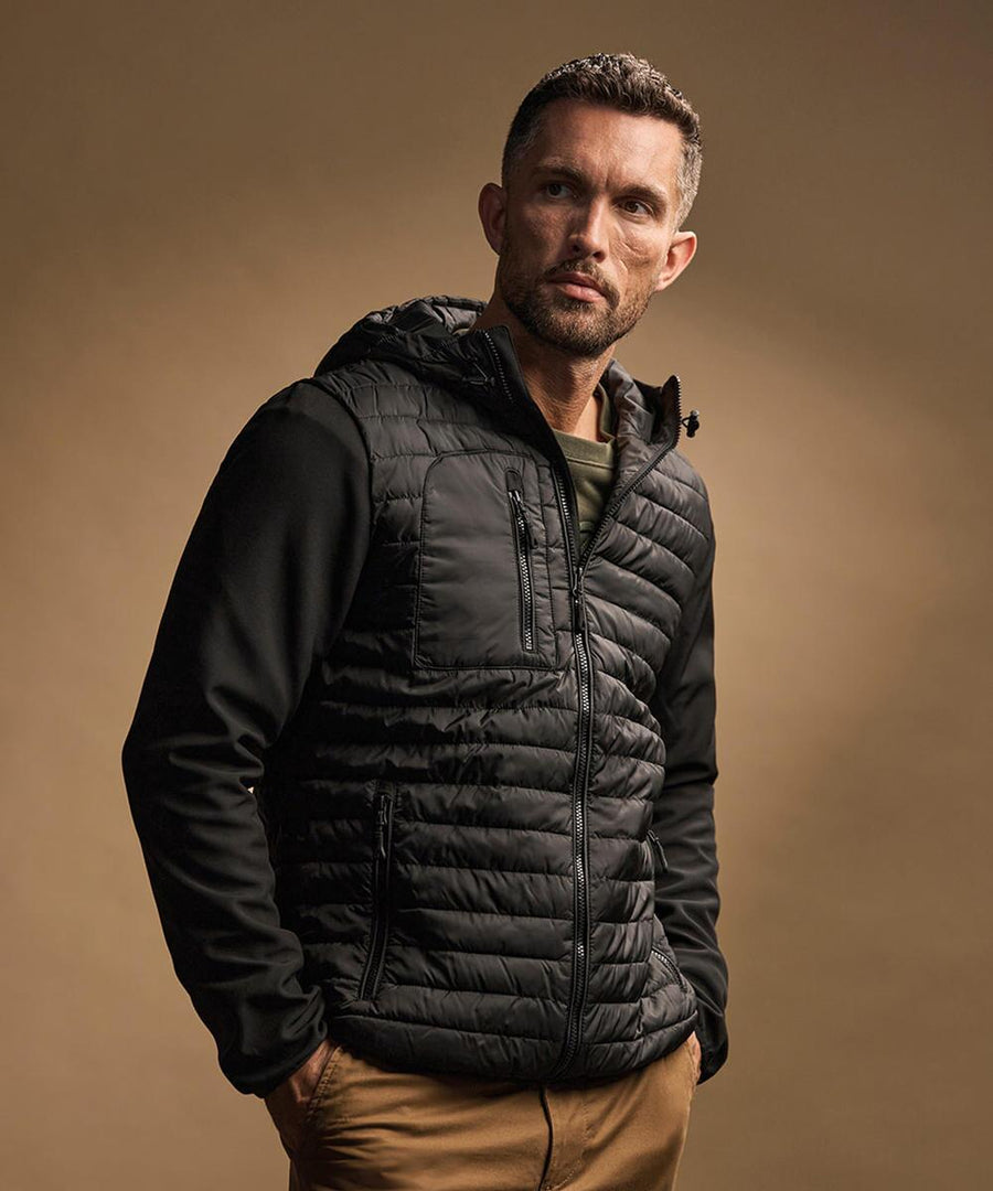 A man with short hair and a trimmed beard wears the Tee Jays Hooded Crossover Jacket (9628) in black over a green shirt and tan trousers, standing against a plain brown background.