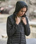 A person with curly hair poses outdoors, wearing the Tee Jays Women's hooded crossover jacket (9629) in black, holding the sides of the hood with both hands against a blurred background.