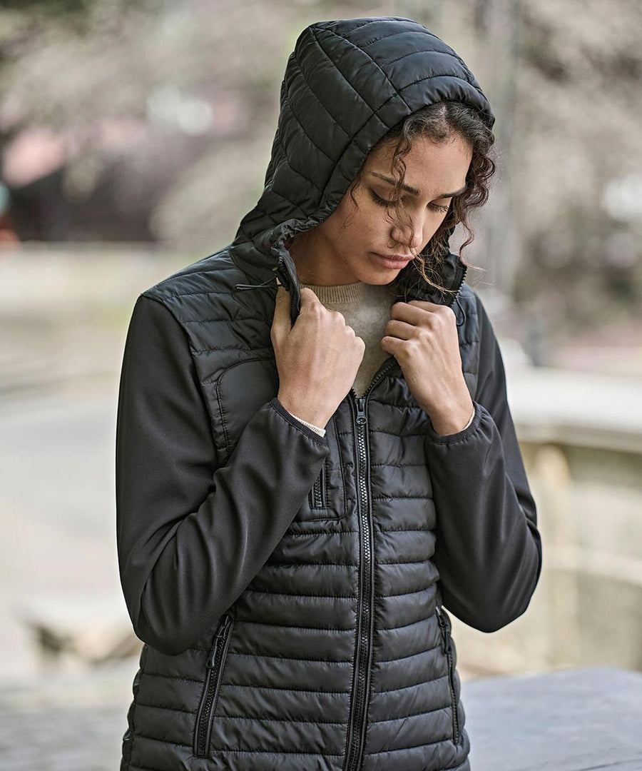 A person with curly hair poses outdoors, wearing the Tee Jays Women's hooded crossover jacket (9629) in black, holding the sides of the hood with both hands against a blurred background.