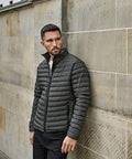 A man with short hair and a beard stands against a stone wall wearing the Tee Jays Zepelin jacket (9630), looking to his left with his hands by his sides.