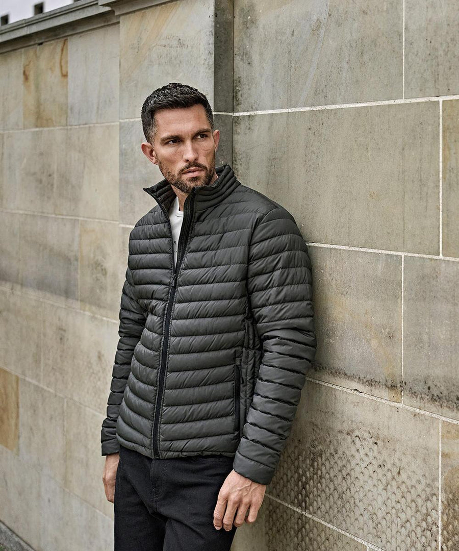 A man with short hair and a beard stands against a stone wall wearing the Tee Jays Zepelin jacket (9630), looking to his left with his hands by his sides.