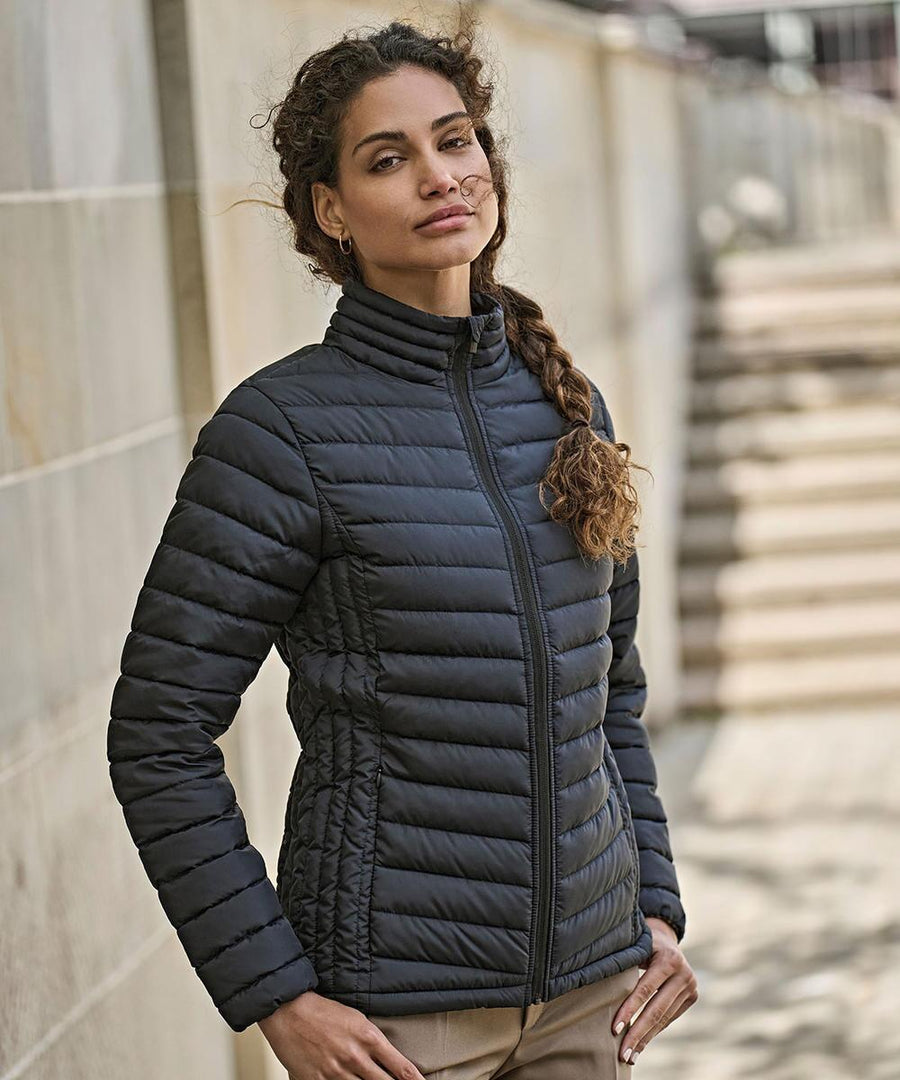 A woman with curly hair in a side plait stands outdoors in front of a stone wall and steps, wearing the black Tee Jays Women’s Zepelin jacket (9631) and beige trousers.