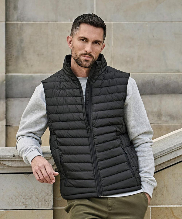 A man with short dark hair and a beard wears the Tee Jays Zepelin bodywarmer (9632), a black quilted gilet, over a light grey sweatshirt and olive trousers as he stands outdoors against a light stone wall, leaning on a curved stone railing.