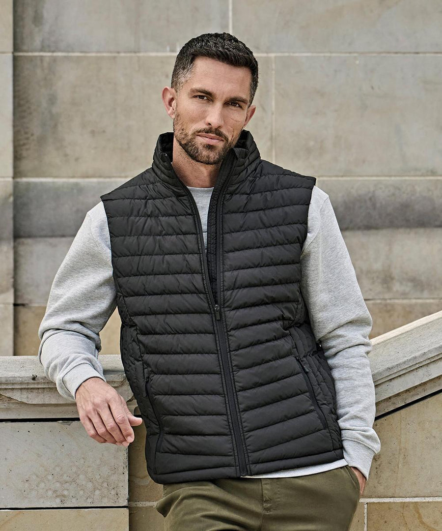 A man with short dark hair and a beard wears the Tee Jays Zepelin bodywarmer (9632), a black quilted gilet, over a light grey sweatshirt and olive trousers as he stands outdoors against a light stone wall, leaning on a curved stone railing.