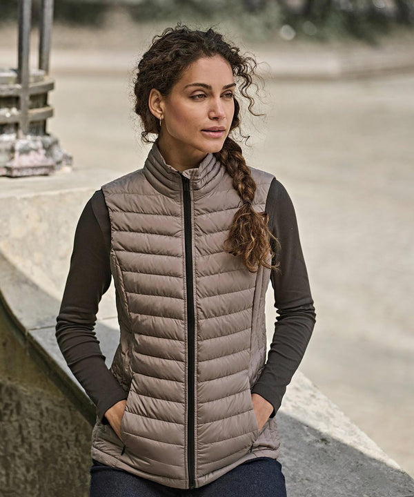 A woman with long, curly brown hair in a plait wears the Tee Jays Womens Zepelin bodywarmer (9633) over a dark long-sleeve shirt, sitting outdoors with hands in the gilet pockets and looking to the side.