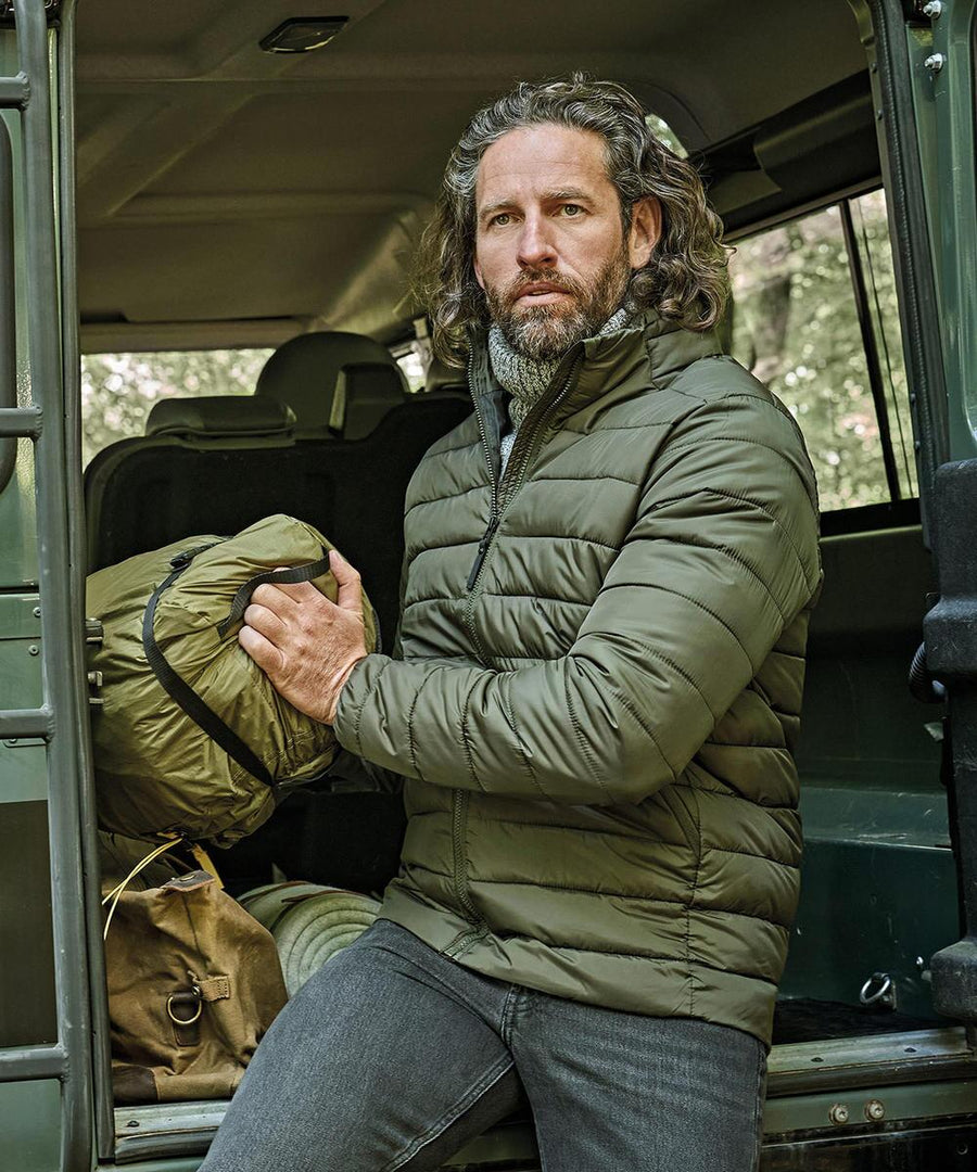 A man with long, wavy hair and a beard sits in the open doorway of a vehicle wearing the Tee Jays Lite jacket (9644) and jeans, holding a green rucksack, with another bag on the vehicle floor next to him.