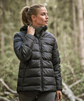 A woman wearing the Tee Jays Women’s Lite Hooded Jacket (9647) and a polo neck stands in a forest, holding the jacket’s collar and looking to the side, with trees and greenery in the background.