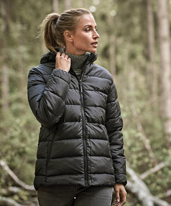 A woman wearing the Tee Jays Women’s Lite Hooded Jacket (9647) and a polo neck stands in a forest, holding the jacket’s collar and looking to the side, with trees and greenery in the background.
