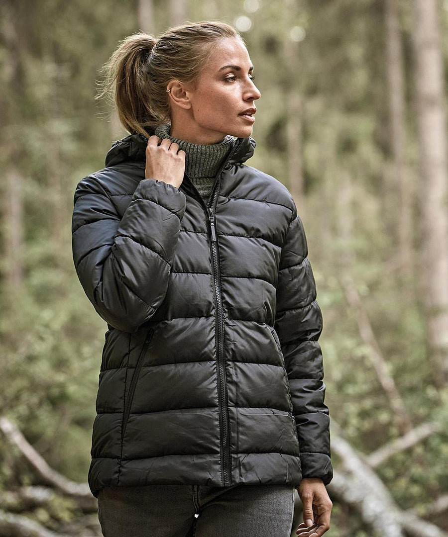 A woman wearing the Tee Jays Women’s Lite Hooded Jacket (9647) and a polo neck stands in a forest, holding the jacket’s collar and looking to the side, with trees and greenery in the background.