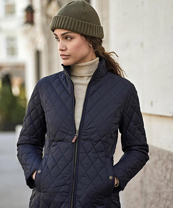 A woman stands outside in an urban setting wearing the Tee Jays Womens Richmond jacket (9661), a quilted navy layer, with her hands in the pockets and looking to the side.