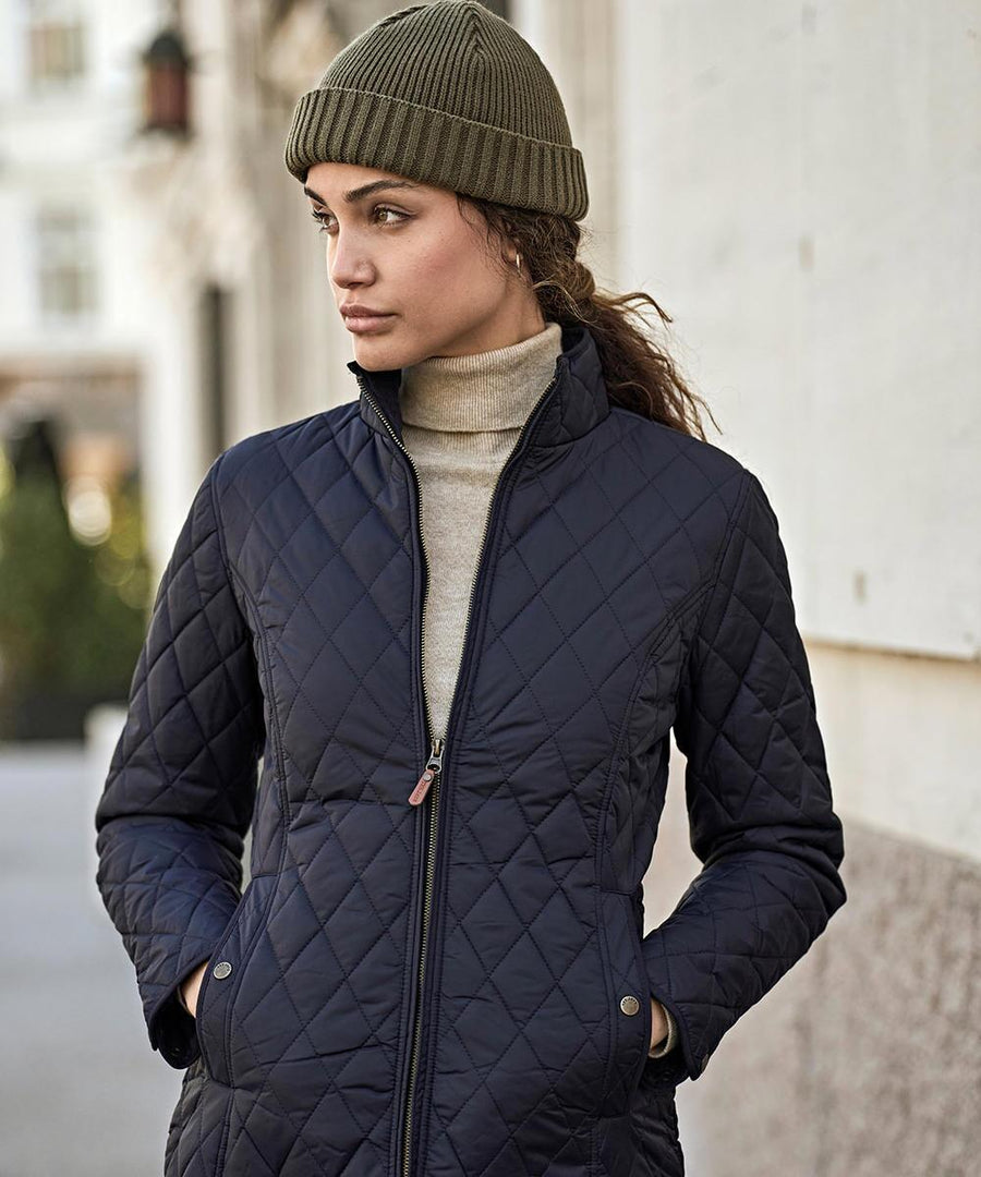A woman stands outside in an urban setting wearing the Tee Jays Womens Richmond jacket (9661), a quilted navy layer, with her hands in the pockets and looking to the side.
