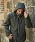 A man wearing the Tee Jays All weather winter jacket (9680) stands against a stone and brick wall, looking down as he adjusts his hood with one hand.