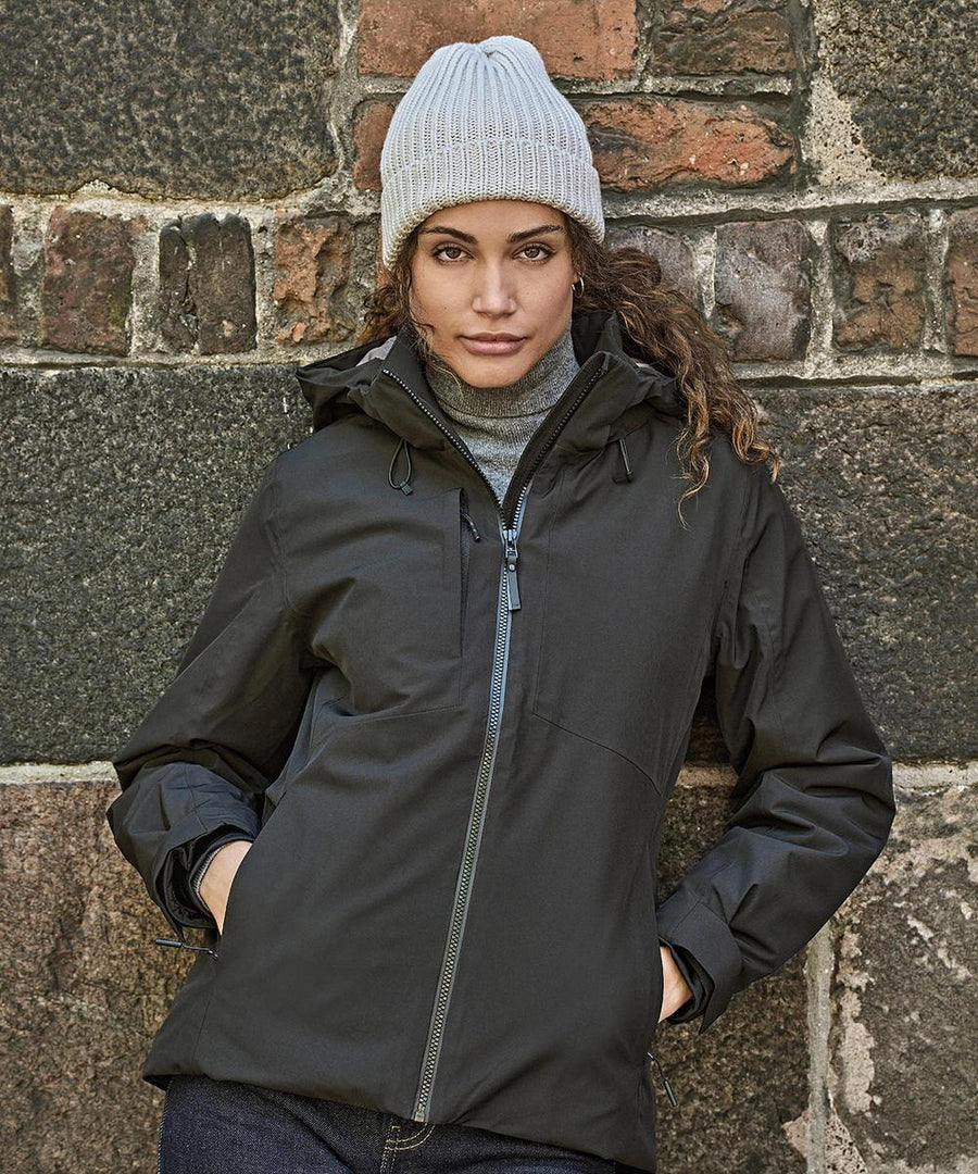 Wearing a grey knit beanie and a grey polo neck, she poses against a stone wall with hands in pockets, showcasing the Tee Jays Womens All Weather Winter Jacket (9681) in dark colour.