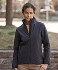 A woman stands outdoors in front of a bench and greenery, wearing the Tee Jays Women’s All Weather Jacket (9607) in navy and light brown trousers, with trees softly blurred in the background.