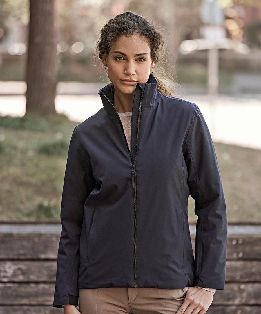 A woman stands outdoors in front of a bench and greenery, wearing the Tee Jays Women’s All Weather Jacket (9607) in navy and light brown trousers, with trees softly blurred in the background.
