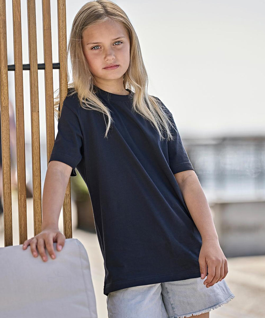 A young girl with long blonde hair wears a Tee Jays Junior UNLABELLED tee (1140B) in navy blue and light denim shorts, standing outdoors while leaning on a wooden structure with one hand on a light-coloured cushion.