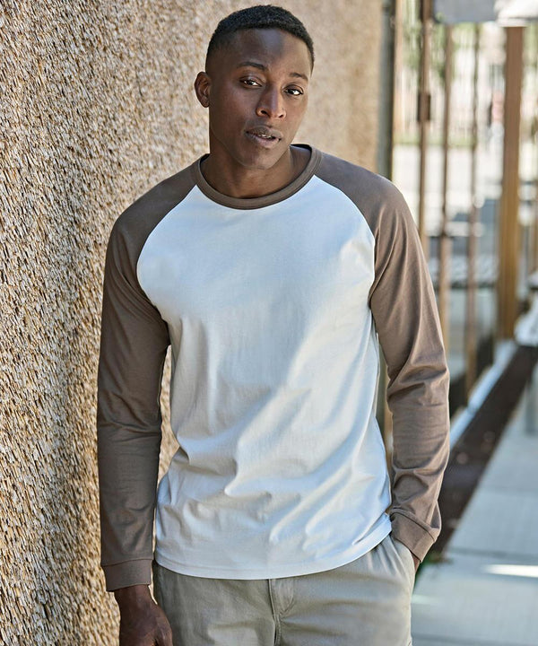 A person stands outdoors by a textured wall, wearing the Tee Jays UNLABELLED baseball tee (1170) with white and brown long sleeves, plus light-coloured trousers, looking at the camera with a neutral expression.