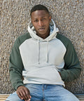 A man sits on a wooden bench in front of a textured wall, wearing the Tee Jays UNLABELED two-tone hooded sweatshirt (5156) with dark green sleeves and blue jeans, looking directly at the camera with a neutral expression.