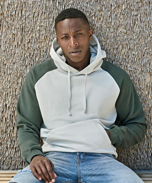 A man sits on a wooden bench in front of a textured wall, wearing the Tee Jays UNLABELED two-tone hooded sweatshirt (5156) with dark green sleeves and blue jeans, looking directly at the camera with a neutral expression.