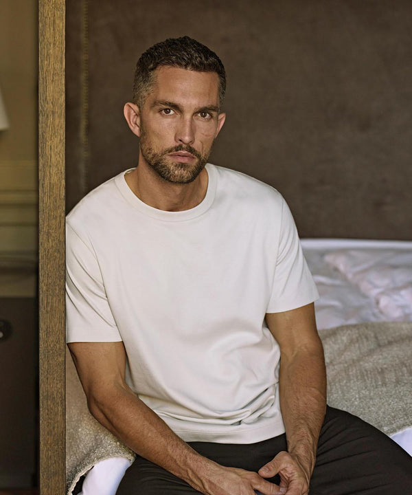 A man with short dark hair and a trimmed beard sits on the edge of a bed, wearing the Tee Jays Pima cotton tee (1450) in white and dark trousers. Behind him are neutral bedding tones and a brown wall.
