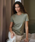 A woman with long curly hair sits indoors on a textured surface, wearing a short-sleeved olive green Women's Pima cotton tee (1451) by Tee Jays and light-coloured trousers, looking at the camera with a neutral expression.