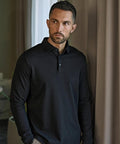 A man with short dark hair and a trimmed beard stands indoors near beige curtains, wearing the Tee Jays Pima cotton long-sleeved polo (1442) and looking slightly to the side with a neutral expression.