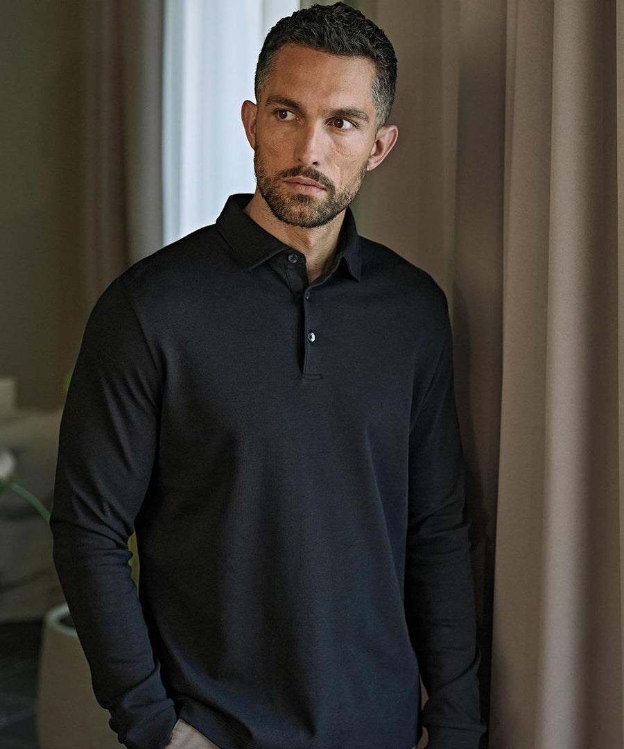 A man with short dark hair and a trimmed beard stands indoors near beige curtains, wearing the Tee Jays Pima cotton long-sleeved polo (1442) and looking slightly to the side with a neutral expression.