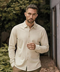 A man with short hair and a beard stands outdoors among green foliage, wearing the Tee Jays Luxury Stretch Shirt (1416) in cream and light brown trousers, looking at the camera with a neutral expression and one hand near his chest.